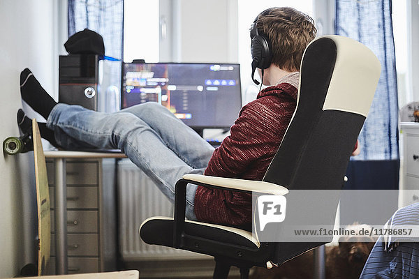 Teenager-Junge sitzt auf einem Stuhl mit den Füßen nach oben am Computer und hört Musik über Kopfhörer zu Hause.