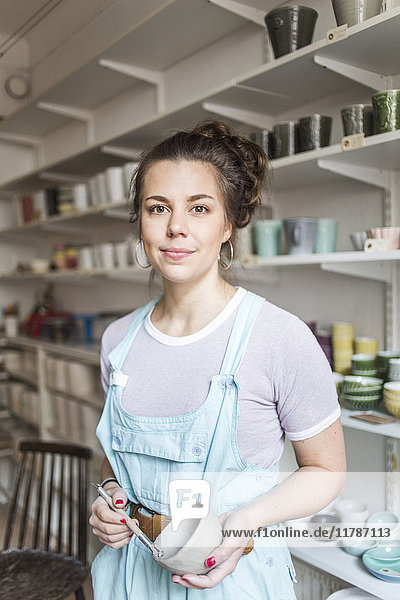 Porträt einer selbstbewussten jungen Töpferin  die Handwerkzeug und Vase hält  während sie im Laden an den Regalen steht.