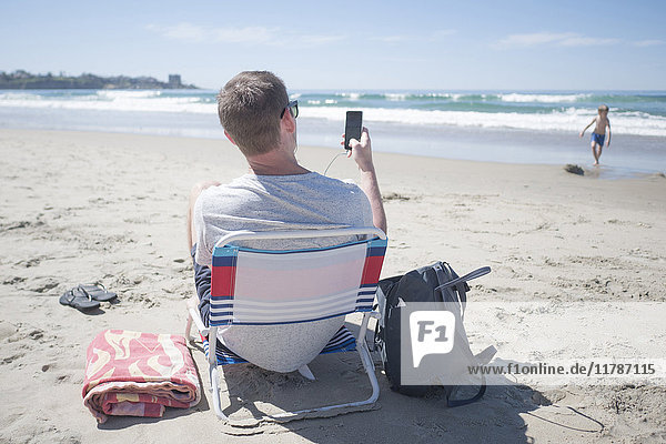 Rear view of man using mobile phone while sitting on chair at beach