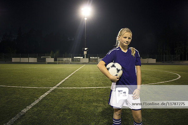 Porträt eines selbstbewussten Mädchens mit Ball auf dem Fußballplatz gegen den Himmel bei Nacht.