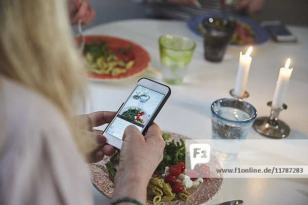 Abgeschnittenes Bild einer Frau  die Essen durch ein Smartphone am Esstisch fotografiert.