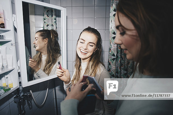 Happy female friends with mobile phone and lip gloss at dorm bathroom