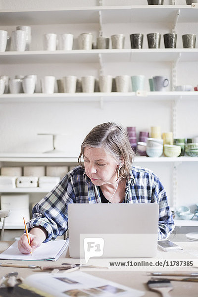 Mature female potter writing on paper while sitting with laptop at table in workshop