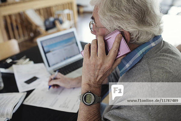 Seitenansicht eines älteren Mannes  der mit dem Handy telefoniert  während er zu Hause Rechnungen prüft.
