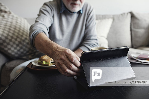 Senior-Mann hält digitale Tablette  während er am Tisch frühstückt