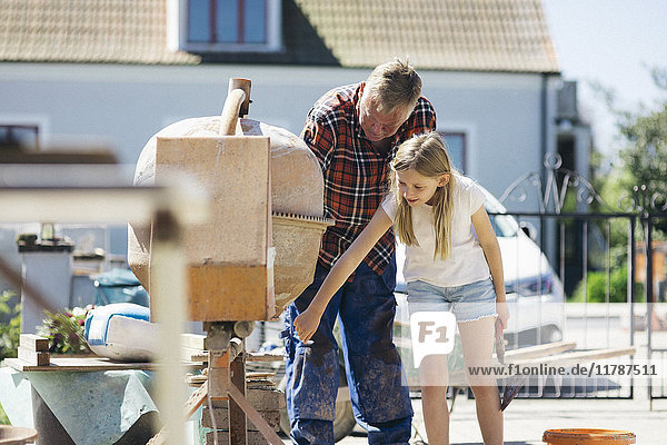 Grandfather using cement mixer with granddaughter in yard