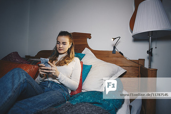 Junge Frau auf dem Bett sitzend mit Handy im Schlafzimmer