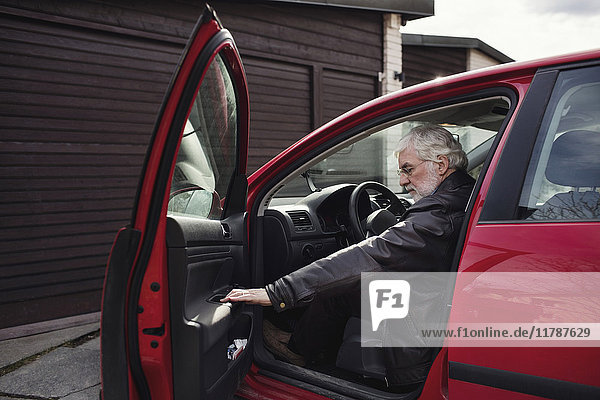 Älterer Mann sitzt in rotem Auto und schließt die Tür gegen die Garage.