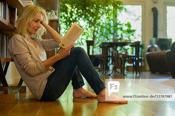 Reife Frau sitzt zu Hause auf dem Boden und liest Buch.