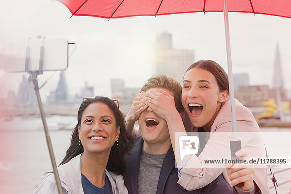 Playful friend tourists with umbrella taking selfie with camera phone selfie stick on bridge  London  UK