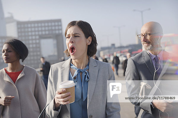 Müde Geschäftsfrau mit Kaffee gähnend auf der sonnigen morgendlichen städtischen Fußgängerbrücke