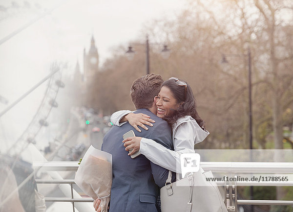 Smiling couple hugging  boyfriend surprising girlfriend with flowers on urban bridge  London  UK