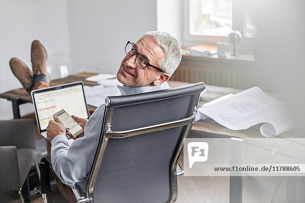 Portrait lächelnder  selbstbewusster Geschäftsmann mit Handy und Laptop im Büro