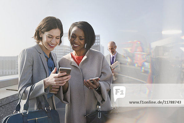 Smiling businesswomen using cell phone on sunny urban bridge