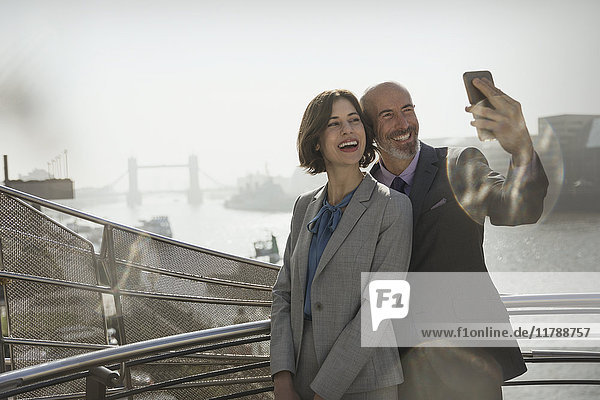 Enthusiastisches  lächelndes Geschäftspaar nimmt Selfie mit Fotohandy auf sonniger urbaner Brücke  London  UK