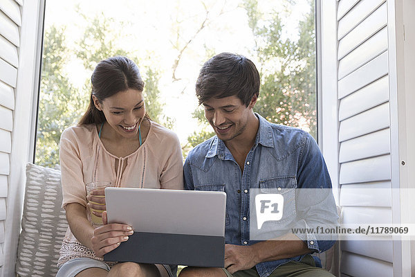 Smiling young couple sitting on windowsill sharing tablet