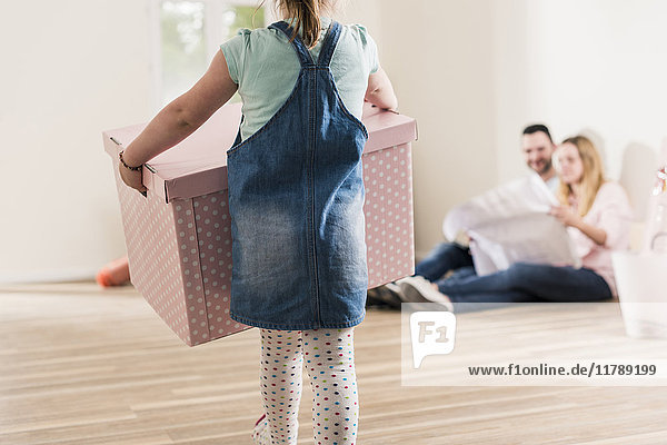 Girl carrying cardboard box in empty apartment with parents in background