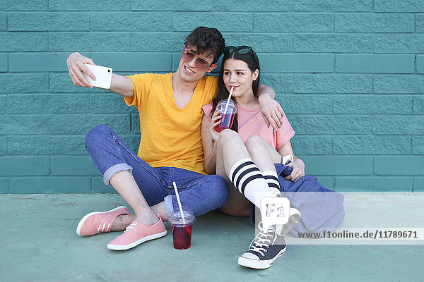 Junges Paar mit Erfrischungsgetränken nimmt Selfie mit Smartphone vor blauer Ziegelwand