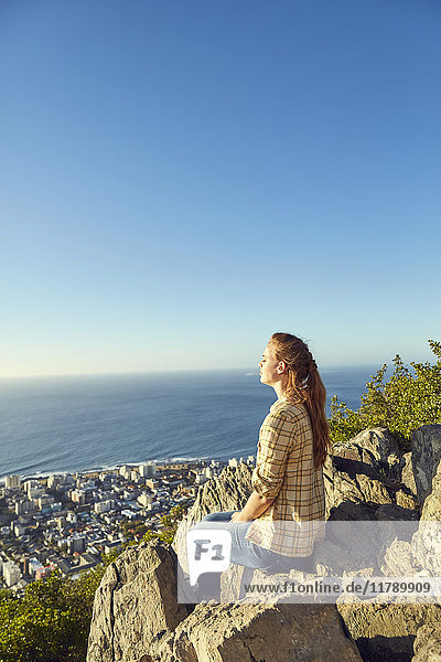 Südafrika  Kapstadt  Signal Hill  junge Frau auf Felsen sitzend mit Blick auf die Stadt und das Meer
