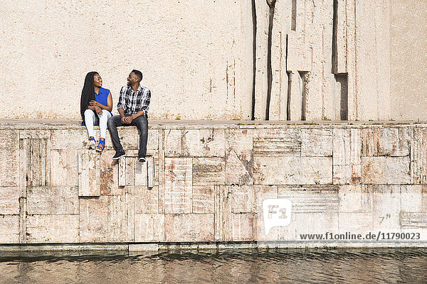 Sprechendes junges Paar auf einer Wand am Wasser sitzend