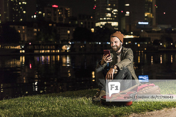 Smiling young man with guitar and cell phone sitting at urban riverside at night