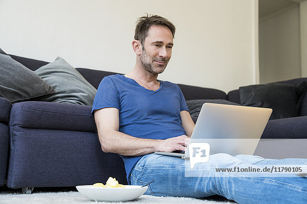 Porträt eines lächelnden Mannes  der im Wohnzimmer mit dem Laptop auf dem Boden sitzt.