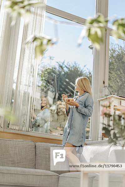 Woman at home standing on couch eating fruit salad looking out of window