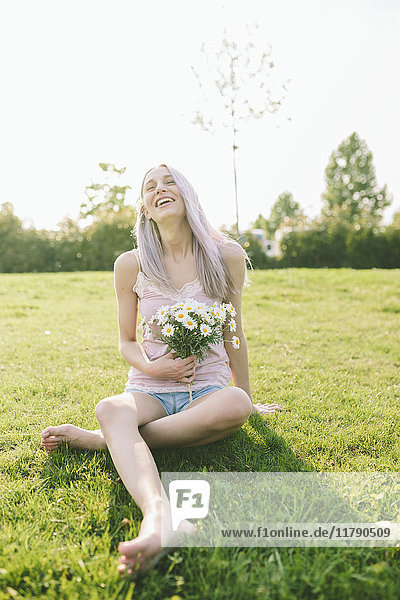 Lachende Frau  die auf einer Wiese sitzt und Gänseblümchen hält.