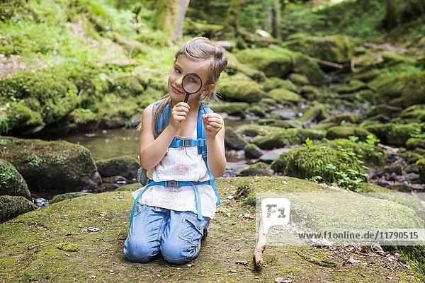 Kleines Mädchen mit Lupe  das im Wald auf einem Felsen hockt und eine Feder beobachtet.