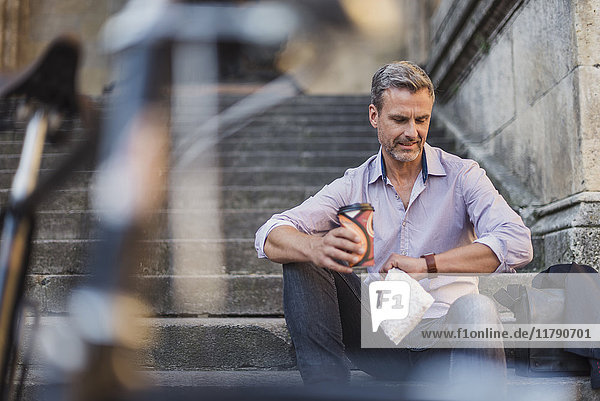 Mann sitzt auf einer Treppe mit Kaffee zum Mitnehmen in der Stadt und überprüft die Uhrzeit.