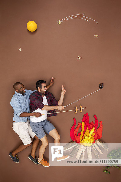 Schwules Paar brät Würstchen am Lagerfeuer und nimmt sich einen Selfie.