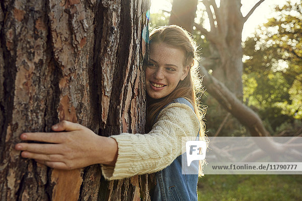 Porträt einer lächelnden jungen Frau  die den Baumstamm im Gegenlicht umarmt.