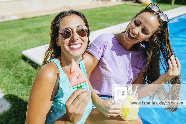 Zwei fröhliche junge Frauen mit Getränk und Wassermelone am Beckenrand