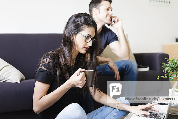 Junge Frau mit Tasse Kaffee mit Laptop im Wohnzimmer  während ihr Freund telefoniert.