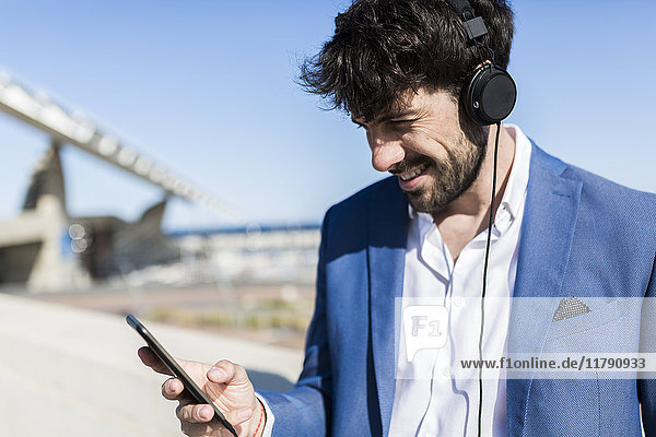 Young businessman wearing headphones looking at smartphone outdoors