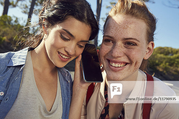 Zwei lächelnde junge Frauen teilen sich ihr Handy im Freien.