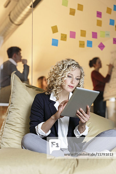 Woman in office using tablet in bean bag with meeting in background