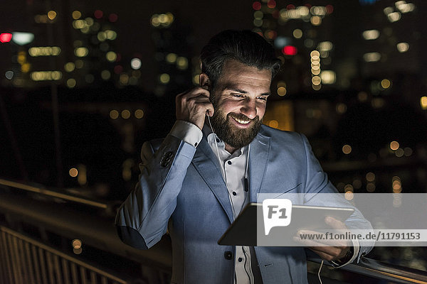 Lächelnder junger Mann mit Tablett und Kopfhörer auf Stadtbrücke bei Nacht
