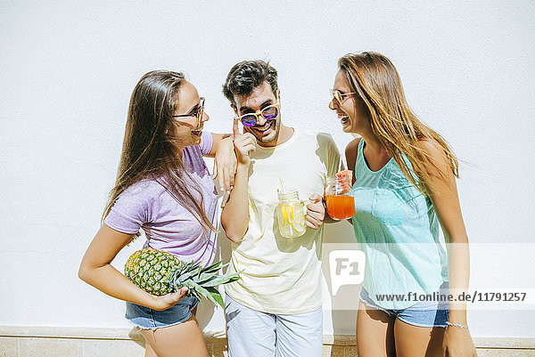 Glückliche Freunde halten erfrischende Getränke und Ananas vor der weißen Wand.