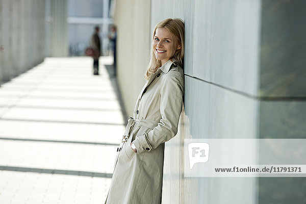 Porträt einer lächelnden blonden Frau im Trenchcoat  die sich an die Fassade lehnt