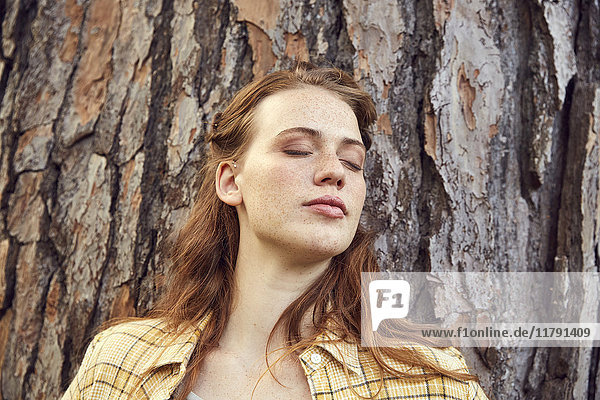 Portrait of redheaded young woman leaning against tree trunk with eyes closed