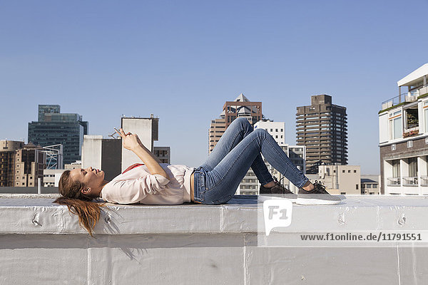 Junge Frau auf Balustrade einer Dachterrasse liegend  mit Smartphone