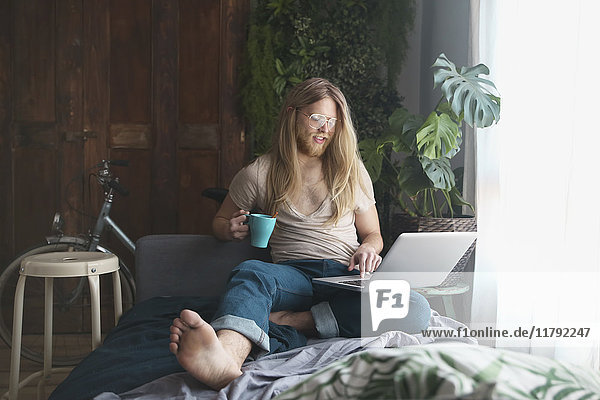 Bärtiger junger Mann mit langen Haaren mit Laptop und Kaffeetrinken zu Hause
