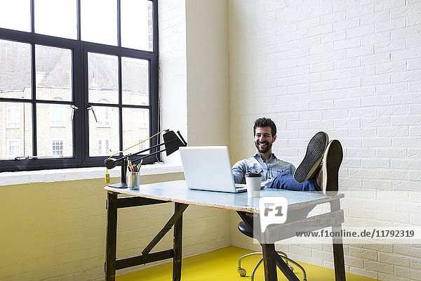 Lächelnder junger Mann sitzt mit erhobenen Füßen am Schreibtisch und schaut auf den Laptop.