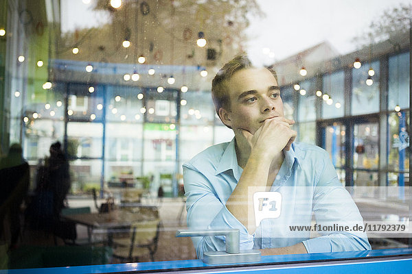 Porträt eines nachdenklichen jungen Mannes  der in einem Café sitzt und durchs Fenster schaut.