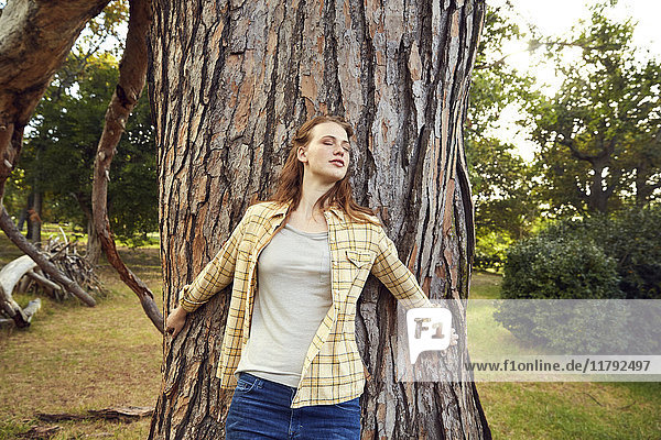 Rothaarige junge Frau lehnt sich mit geschlossenen Augen an den Baumstamm.