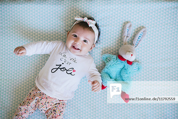 Happy baby girl lying on bed beside toy bunny