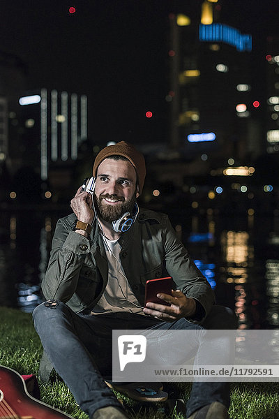 Lächelnder junger Mann mit Gitarre  Handy und Kopfhörer sitzt nachts am städtischen Flussufer.