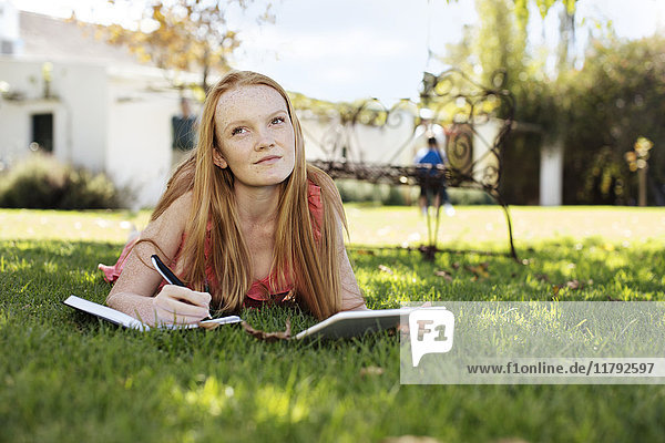Grl with long red hair lying in grass with tablet and notebook