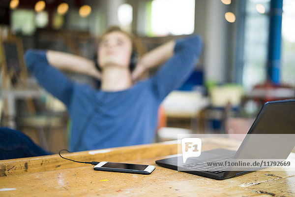 Laptop und Smartphone auf dem Tisch mit entspannendem jungen Mann  der im Hintergrund Musik hört.